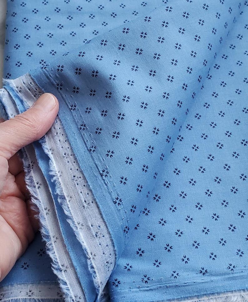 May include: A close-up of blue fabric with a repeating pattern of small, dark blue floral shapes. The fabric has a light blue background and a frayed edge. The fabric is folded. The image shows the edge of the fabric.