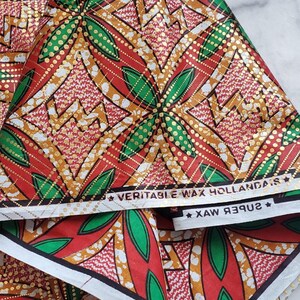 May include: A folded piece of fabric featuring a vibrant geometric pattern in red, green, and gold. The design includes repeating diamond shapes and floral motifs with gold dotted accents. The white selvage edge displays the text VERITABLE WAX HOLLANDAIS.