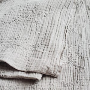 以下が含まれることがあります： 繊細な同系色の花柄刺繍が施された、ライトベージュの多層モスリン生地。生地全体にシワ感のあるテクスチャーがあり、縦方向のステッチラインと柔らかく折り畳まれたエッジが特徴的です。