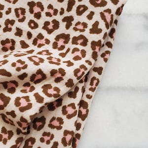 Könnte beinhalten: Gefalteter Stoff mit beigem Hintergrund und einem sich wiederholenden Muster aus braunen und rosa Leopardenflecken. Der Stoff ist weich und wirkt leicht. Das Leopardenmuster ist ein klassisches und vielseitiges Muster.