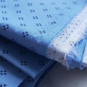 May include: Folded blue fabric with a repeating pattern of small, dark blue floral shapes. The fabric has a light blue base with a white border and a frayed edge, suggesting a textile or craft material. The image is a close-up.