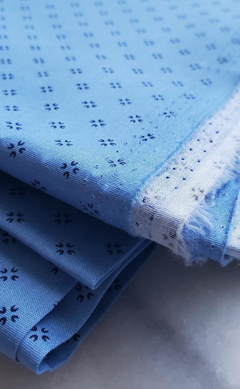 May include: Folded blue fabric with a repeating pattern of small, dark blue floral shapes. The fabric has a light blue base with a white border and a frayed edge, suggesting a textile or craft material. The image is a close-up.