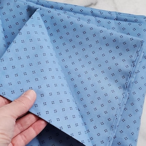 May include: Pale blue fabric featuring a repeating pattern of tiny, dark blue floral motifs. The fabric is folded, with a hand holding a section of the material. The fabric appears to be cotton or a similar material.