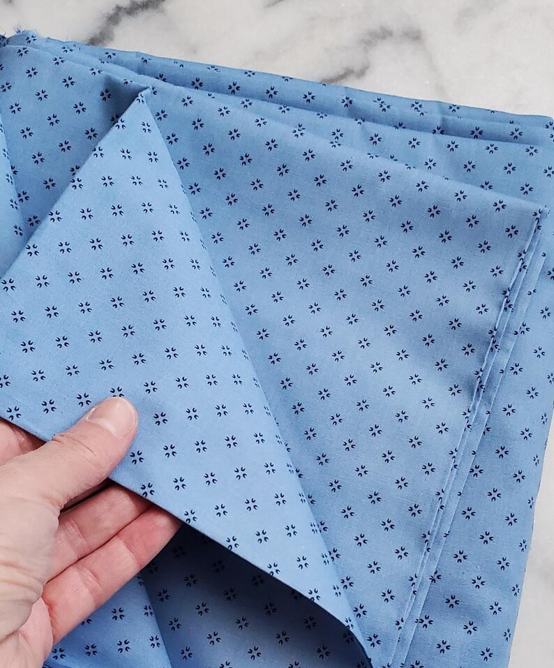 May include: Pale blue fabric featuring a repeating pattern of tiny, dark blue floral motifs. The fabric is folded, with a hand holding a section of the material. The fabric appears to be cotton or a similar material.