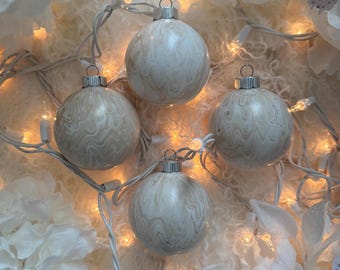 May include: Four white and beige swirl-patterned Christmas ornaments with silver caps are arranged on a white, textured surface. A string of warm white lights weaves between the ornaments, and white floral accents surround them.