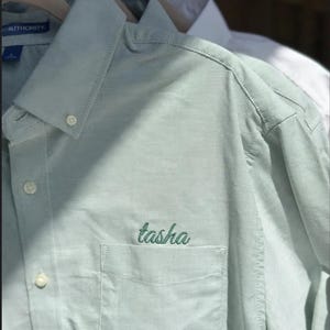 May include: Light green button-down shirt with a chest pocket. The name "tasha" is embroidered in green thread on the pocket. The shirt is hanging on a wooden hanger with other shirts in the background.