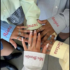 May include: Five hands joined in a circle, each wearing a long-sleeved shirt with a cuff embroidered in red thread. The names Alana, Imani, Amoyea, Clarissa, and Mrs. Brookins are stitched onto the cuffs of light blue, green, grey, yellow, and striped shirts.