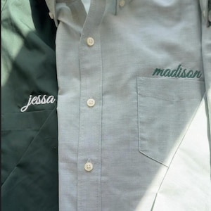 May include: A light blue button-down shirt with a chest pocket embroidered with the name madison in dark green script, positioned next to a dark green shirt featuring the name jessa embroidered in white script on the chest pocket area.