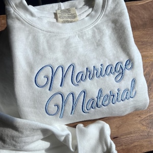 May include: White sweatshirt with blue embroidered text that reads "Marriage Material".