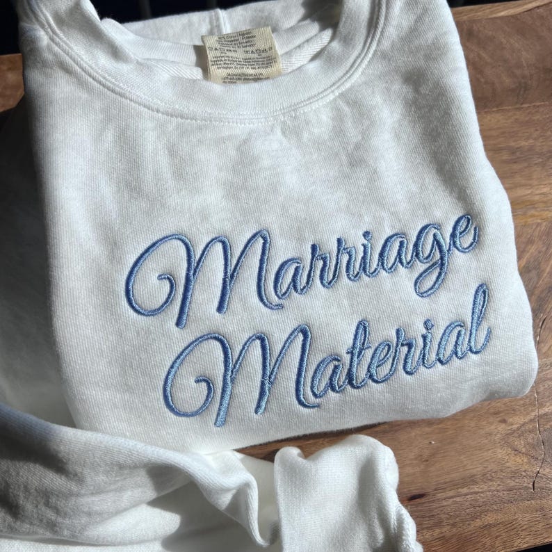 May include: White sweatshirt with blue embroidered text that reads "Marriage Material".