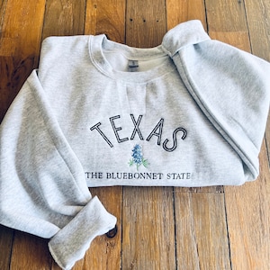 May include: A gray sweatshirt with the word "TEXAS" embroidered in black outlined letters. Below the word is a bluebonnet flower and the text "THE BLUEBONNET STATE".