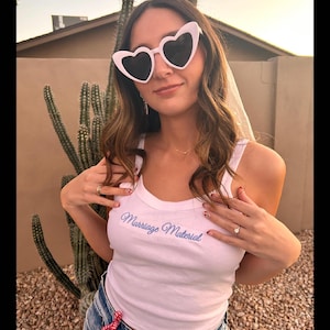 May include: A white tank top with the phrase "Marriage Material" in light blue script. The person is wearing heart-shaped sunglasses with black lenses. The person is also wearing blue jeans.