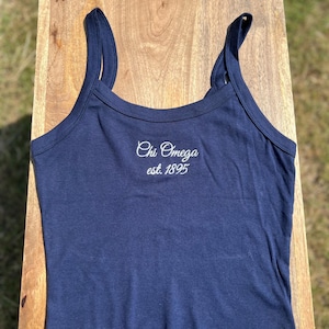 May include: A navy blue tank top with thin straps. The text "Chi Omega est. 1895" is embroidered in white script across the chest. The top is made of a soft, stretchy material and is likely for casual wear.