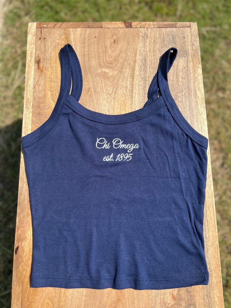 May include: A navy blue tank top with thin straps. The text "Chi Omega est. 1895" is embroidered in white script across the chest. The top is made of a soft, stretchy material and is likely for casual wear.