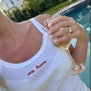May include: White tank top with red embroidered text "mrs. travers". A hand holds a wine glass filled with a light-colored liquid. A large diamond ring is visible on the ring finger. The background includes a pool.