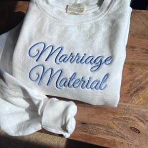 May include: White sweatshirt with blue embroidered text that reads "Marriage Material".
