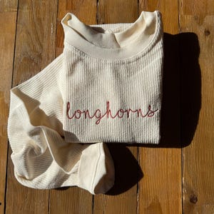 May include: Cream-colored waffle-knit long-sleeved shirt with the word "longhorns" embroidered in red script. The shirt is folded neatly on a wooden surface, with the sleeve partially visible.