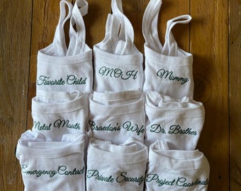 May include: Nine white tank tops with green embroidered text. The text on the tank tops reads: "Favorite Child", "MOH", "Mommy", "Metal Mouth", "Braedan's Wife", "Dr. Brother", "Emergency Contact", "Private Security", and "Project Coordinator".