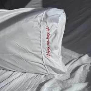 May include: A white pillowcase with the embroidered text "Happy wife, happy life!" in red script. The pillowcase is made of a smooth, possibly satin-like material and is resting on a white sheet.