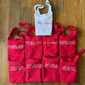 May include: A collection of red and white tank tops with various phrases embroidered in white. The phrases include "Certified Glizzy Connoisseur," "Perfect Tits & Heart," "I'm a Mommy," and "Catch Flights, Not Feelings." One white tank top reads "Mrs. French" in red.