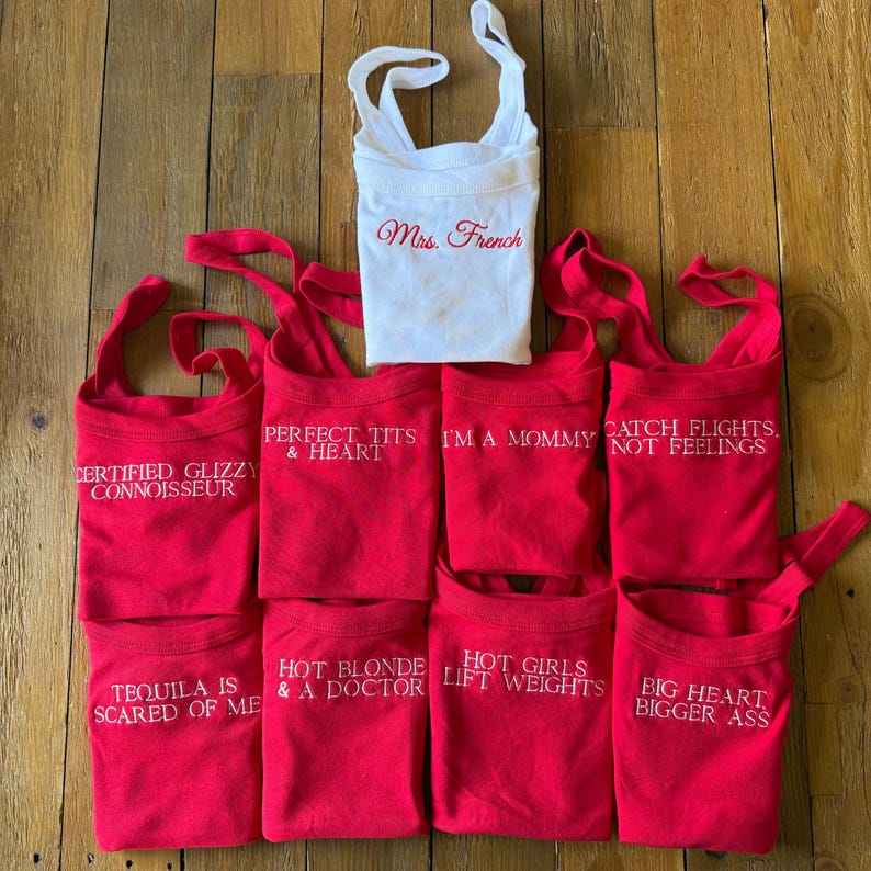 May include: A collection of red and white tank tops with various phrases embroidered in white. The phrases include "Certified Glizzy Connoisseur," "Perfect Tits & Heart," "I'm a Mommy," and "Catch Flights, Not Feelings." One white tank top reads "Mrs. French" in red.