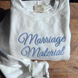 May include: White sweatshirt with blue embroidered text that reads "Marriage Material".