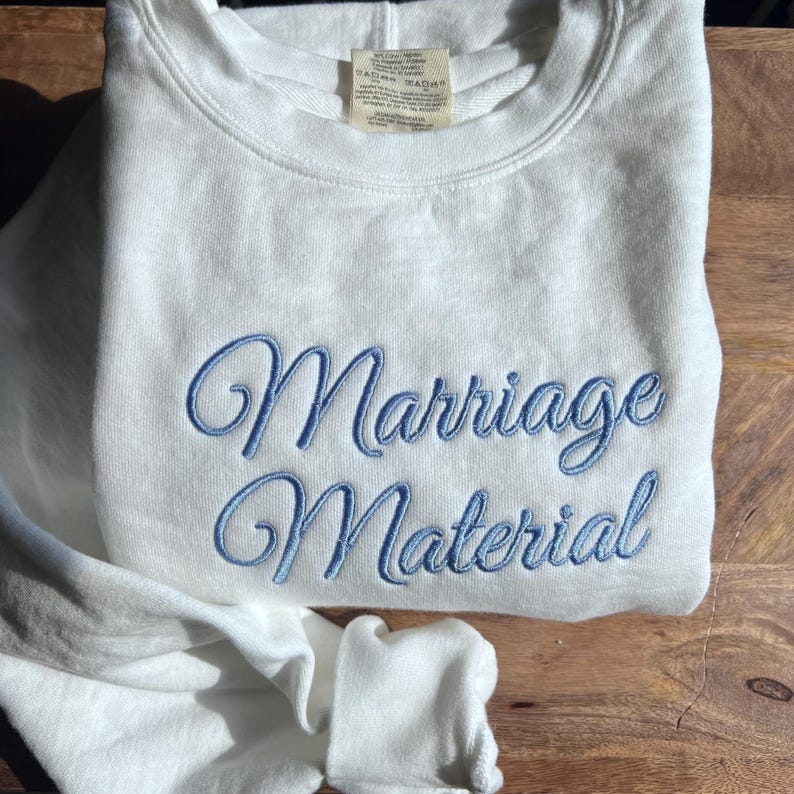May include: White sweatshirt with blue embroidered text that reads "Marriage Material".