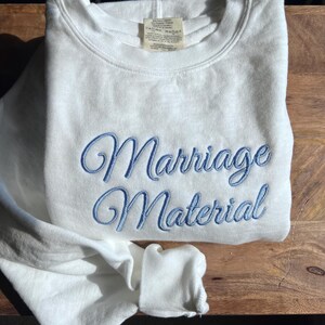 May include: White sweatshirt with blue embroidered text that reads "Marriage Material".