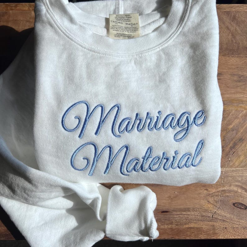 May include: White sweatshirt with blue embroidered text that reads "Marriage Material".