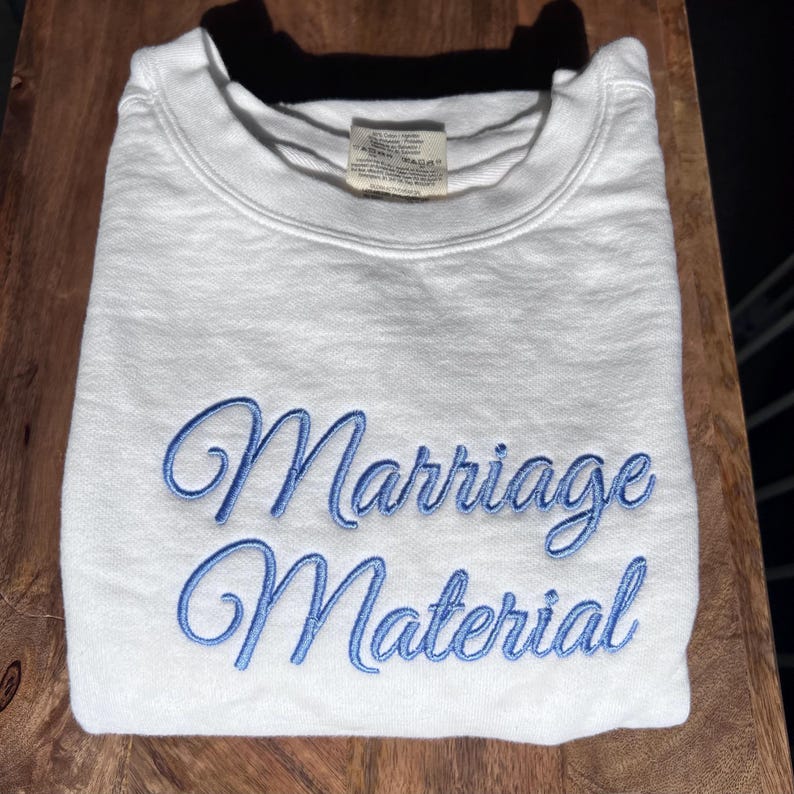 May include: White sweatshirt with blue embroidered text that reads "Marriage Material".