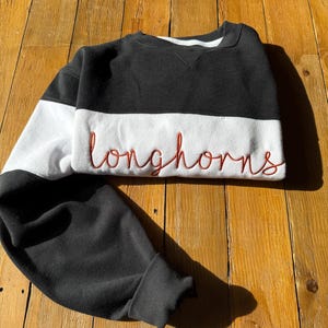 May include: A black, white, and red sweatshirt with the word "longhorns" embroidered in red script across the chest. The sweatshirt has a crew neck and long sleeves. The sleeves and upper portion of the sweatshirt are black, with a white band across the chest.
