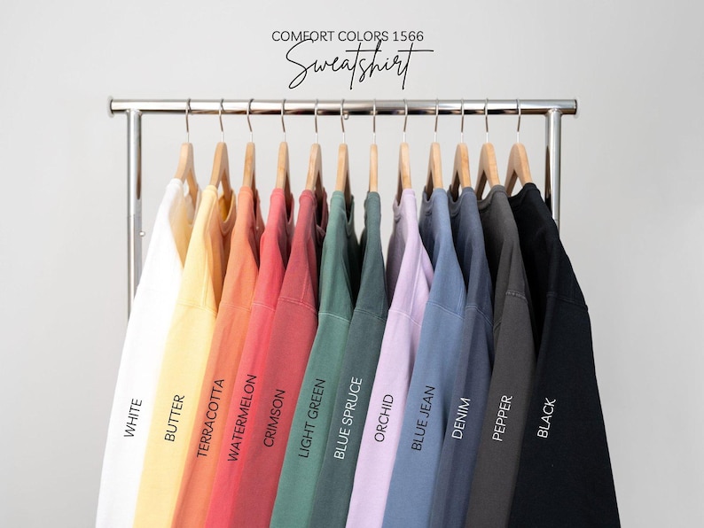 May include: A rack of 12 sweatshirts in various colors, including white, butter, terracotta, watermelon, crimson, light green, blue spruce, orchid, blue jean, denim, pepper, and black. The sweatshirts are labeled with the color names below each one. The text "COMFORT COLORS 1566 Sweatshirt" is at the top of the image.
