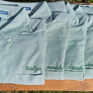 May include: A set of light blue button-down shirts with long sleeves, a collar, and a chest pocket. Each shirt features the embroidered name tristyn, maddie, brooke, shania, or sunny in dark green cursive script. The label reads Port Authority.