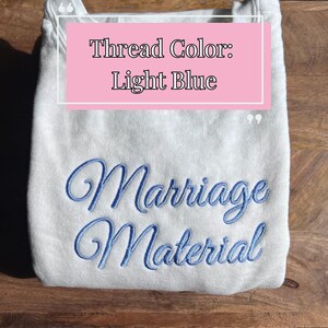 May include: A light gray sweatshirt with the text "Marriage Material" embroidered in light blue thread. The text "Thread Color: Light Blue" is written in pink on a white background.