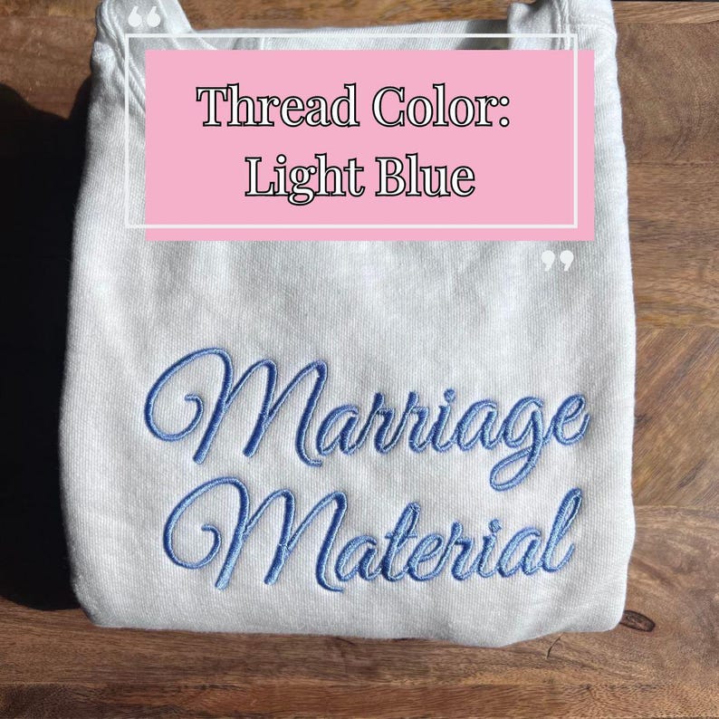 May include: A light gray sweatshirt with the text "Marriage Material" embroidered in light blue thread. The text "Thread Color: Light Blue" is written in pink on a white background.