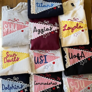 May include: A collection of sweatshirts featuring embroidered pennant designs with lace trim. Each pennant displays team names like Titans, SHU, Aggies, Loyola, Sun Devils, USI, UofA, Dolphins, Commodores, and SJU, accented with small hearts and stars.