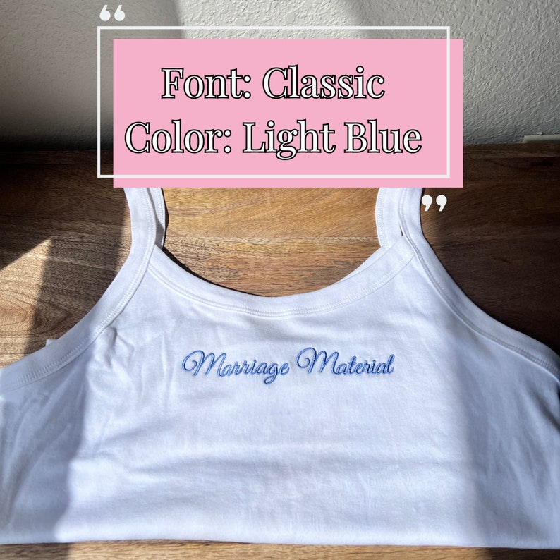 May include: White tank top with the embroidered text "Marriage Material" in light blue script. The image also includes text that says "Font: Classic Color: Light Blue". The tank top is on a wooden surface.