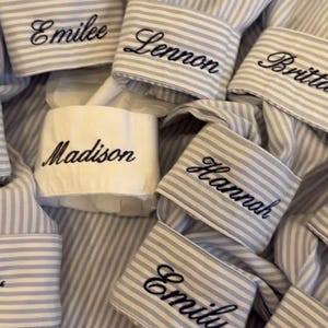 May include: A collection of light blue and white striped shirt cuffs, each featuring black embroidered names including Emilee, Lennon, Britt, Madison, Hannah, and Emily. One cuff is solid white with the name Madison embroidered in black.