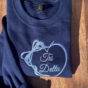 May include: A navy blue sweatshirt with a light blue embroidered heart design and the words "Tri Delta". The heart is surrounded by a bow. The sweatshirt is folded on a wooden surface.