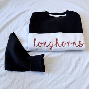 May include: A black and white sweatshirt with the word "longhorns" embroidered in a cursive, reddish-brown font. The sweatshirt is folded neatly on a white surface, with one sleeve folded over.
