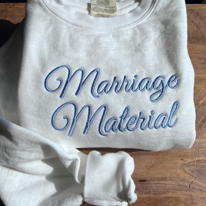 May include: White sweatshirt with blue embroidered text that reads "Marriage Material".