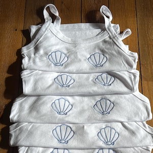 May include: A stack of five white tank tops with blue embroidered seashells on each top. The shells are arranged in a symmetrical pattern.