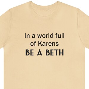 Könnte beinhalten: Cremefarbenes T-Shirt mit dem schwarzen Text: "In a world full of Karens BE A BETH". Das T-Shirt hat einen Rundhalsausschnitt und kurze Ärmel. Der Text ist mittig auf der Vorderseite des Shirts platziert.