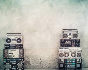 May include: A stack of four vintage boomboxes in black and silver with a textured gray background. The boomboxes have cassette and radio features.