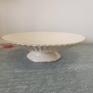 May include: A cream-colored ceramic pedestal bowl featuring a shallow, wide basin with a fluted, scalloped edge. The rim of the bowl and the base of the pedestal are accented with a thin, delicate gold trim, providing a refined finish to the piece.