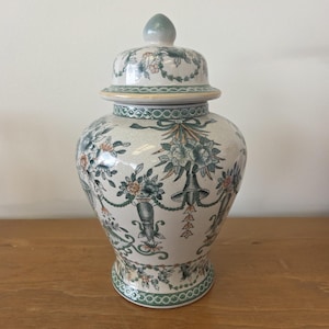 May include: A ceramic ginger jar with a removable lid, featuring a cream-colored base with a crackle glaze finish. The surface is decorated with a repeating pattern of green and peach-colored floral arrangements, urns, and bows in a neoclassical style.