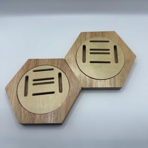 Peut inclure: Deux dessous de verre hexagonaux en bois avec une incrustation circulaire en bois clair. Chaque incrustation comporte trois fentes horizontales et deux fentes verticales, formant un motif en grille. Le grain du bois est visible sur les cadres.