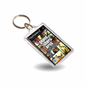 May include: A rectangular keychain featuring the Grand Theft Auto: San Andreas game cover art. The keychain has a clear, plastic casing with a metal ring for attaching keys. The PlayStation 2 logo is visible.