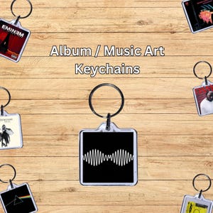 May include: A collection of Album / Music Art Keychains, each with a different design, are shown against a wooden backdrop. The keychains feature album art and music-related designs. One keychain has a black background with a white sound wave.