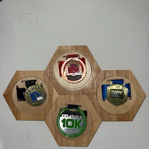 Puede incluir: Un expositor de medallas de madera con cuatro huecos hexagonales, cada uno con una medalla. Las medallas son de varios colores, incluyendo oro, bronce y verde, con cintas y texto. El expositor es de madera clara.